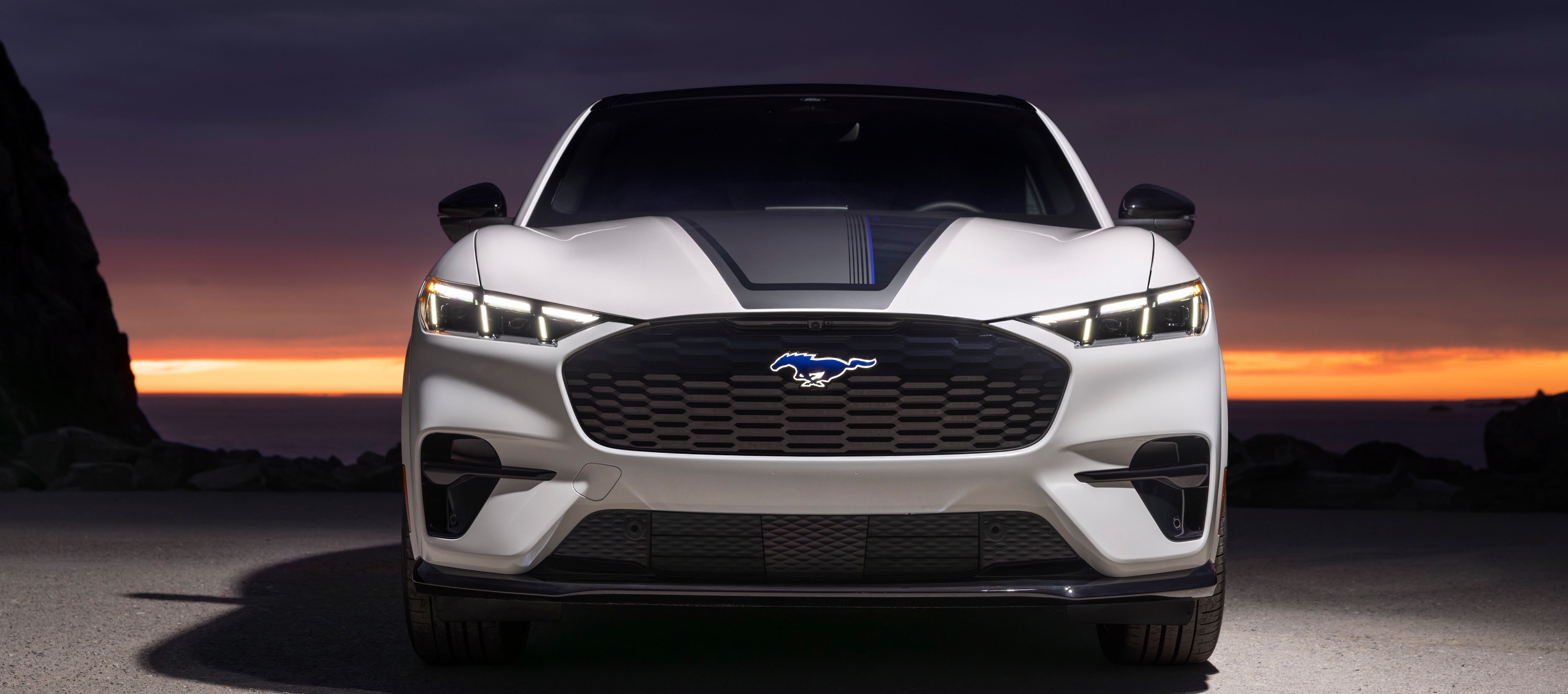 A white Mustang Mach-e is parked in front of a bright orange sunset.